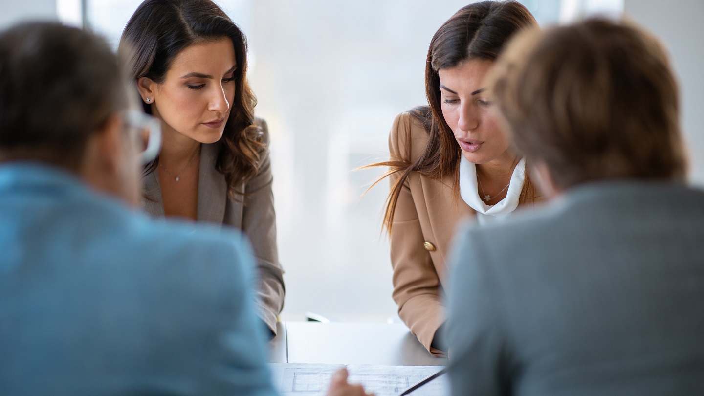 Two businesswomen sitting in a joint meeting with partners and discussing details on blueprint