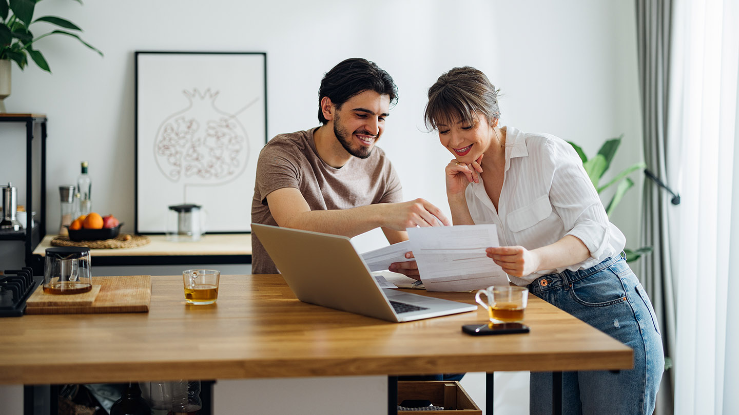 Cheerful boyfriend and girlfriend laughing while doing home finances together online on a laptop computer in the kitchen.